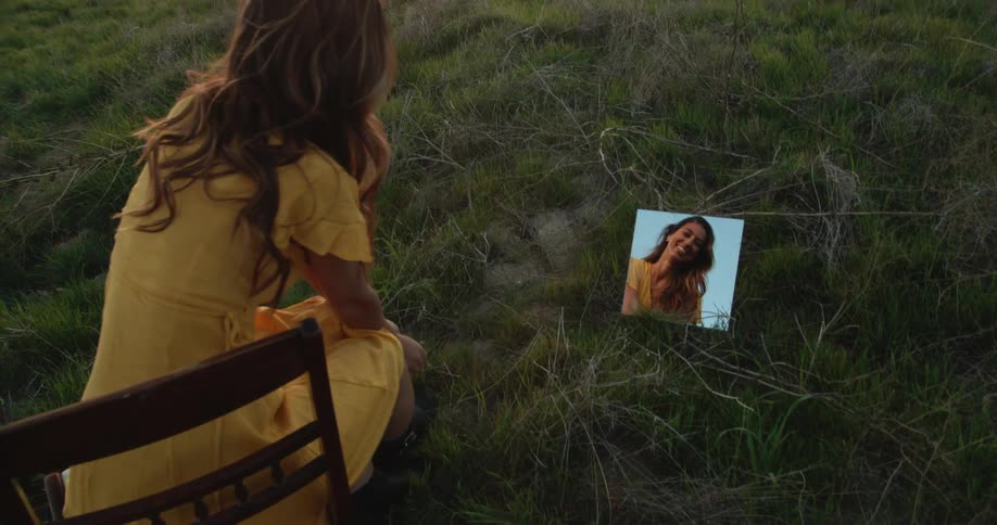 Woman with brown wavy hair and yellow dress sits in chair and looks down in small mirror smiling in the grassy field