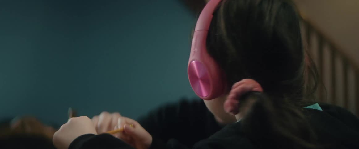 A young girl wears headphones while doing homework on a laptop with her father