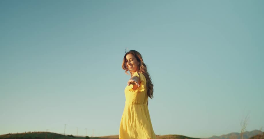 Woman wearing mid thigh long yellow flowing dress walks away reaching and beckoning with hills in background on sunny clear day