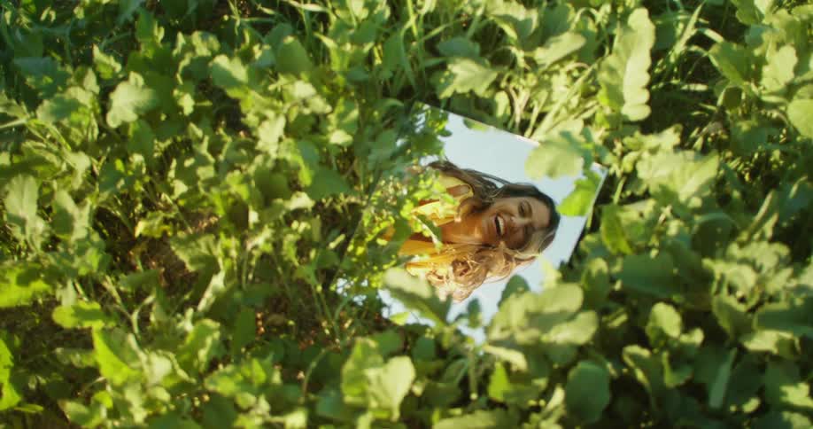 A young woman smiles into a small mirror lying in the grass on a clear day