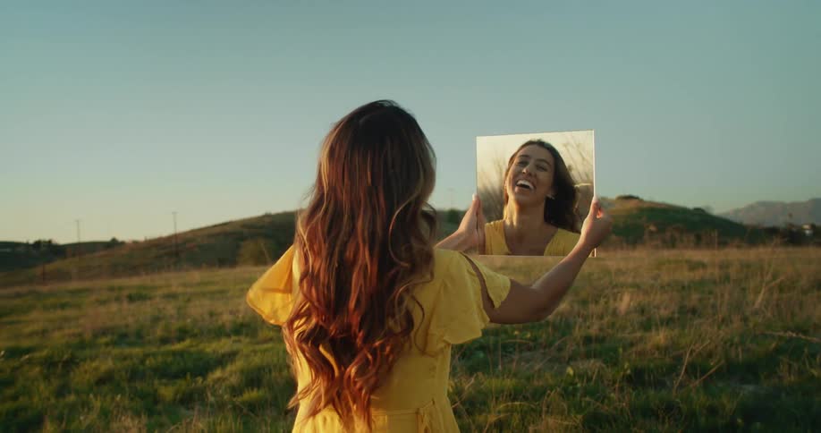 Young woman wearing a yellow dress holds up a mirror and smiles in a field at sunset