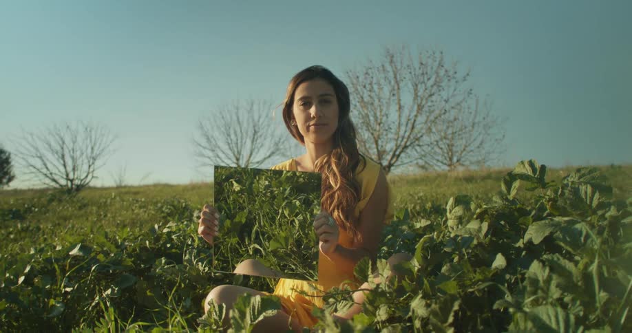 Woman with brown wavy hair and yellow dress holds a mirror sitting in green grass in the grassy field