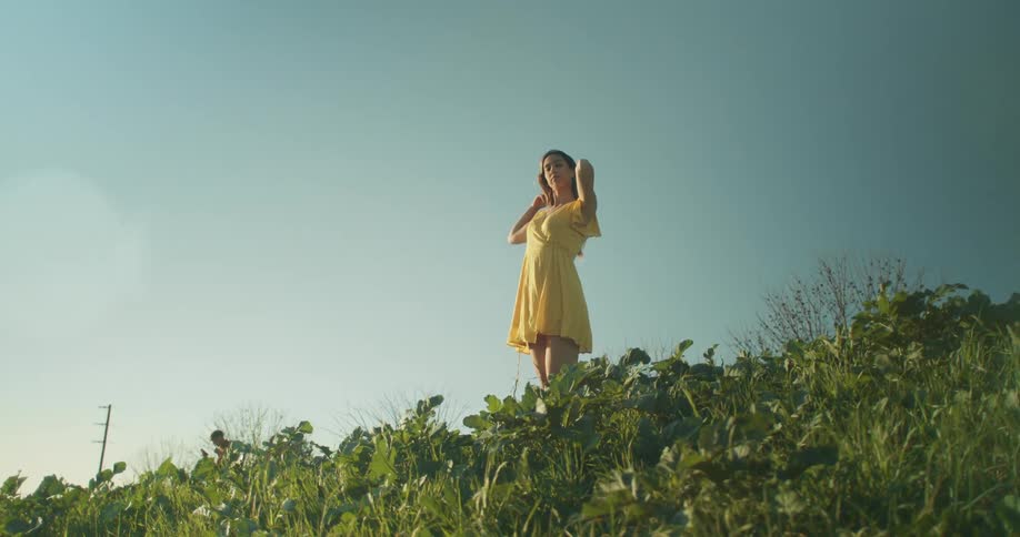 Woman wearing yellow dress stands at top of hill under clear blue sky and combs hair back