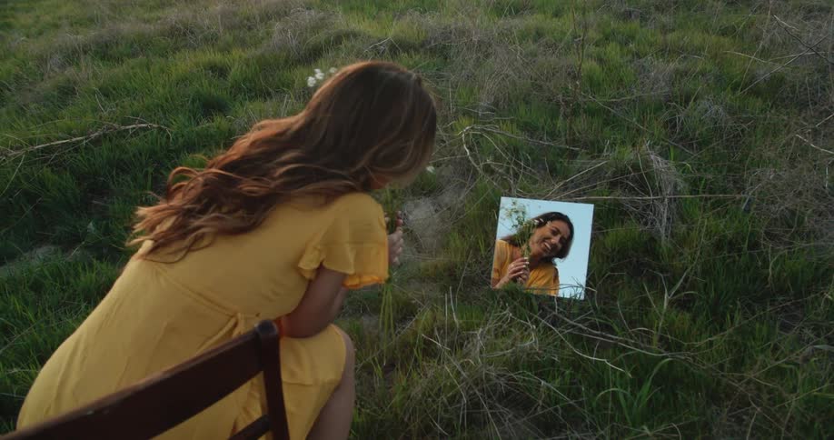 A woman holding wild flowers outdoors looks at a small mirror and laughs