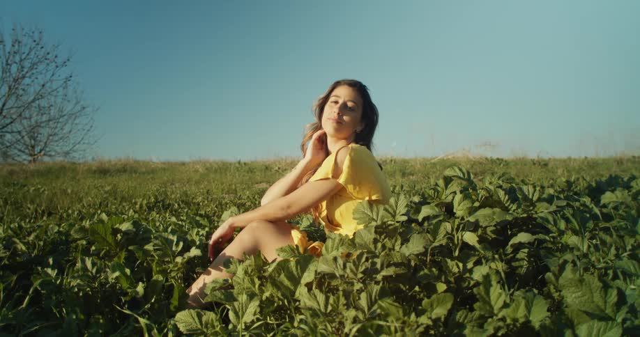 Woman with brown wavy hairand yellow dress sits in grass and looks at the camera in the grassy field
