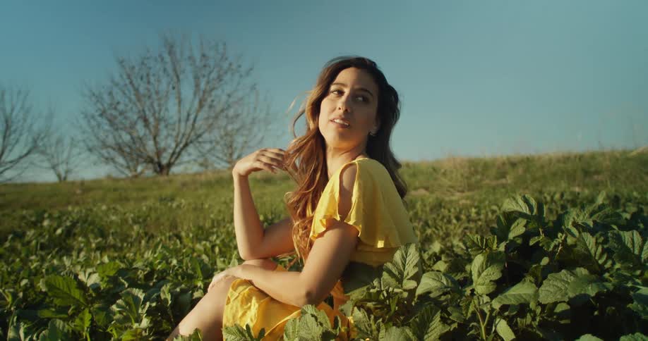 Woman in yellow dress sits in plant patch on grassy slope smiling