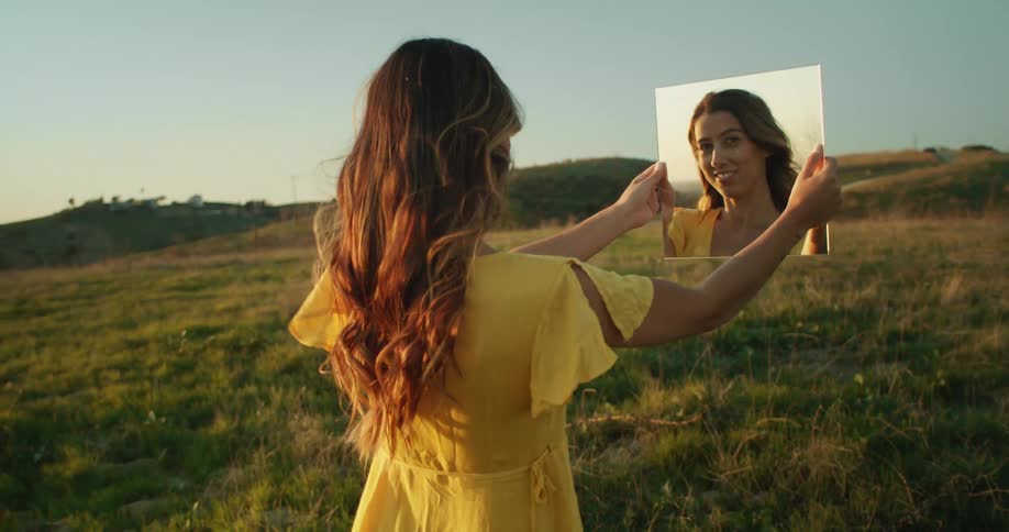 Woman wearing a yellow dress standing on a field looks at the camera using a mirror