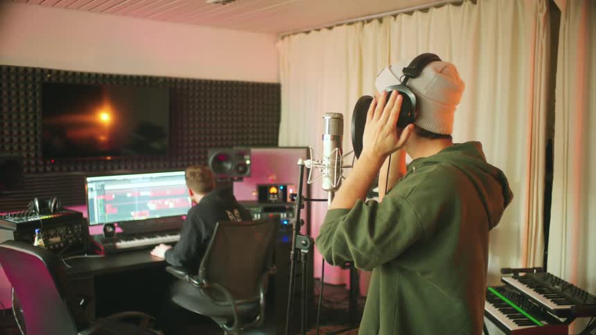 A man is singing into a microphone while another person is monitoring on a computer nearby