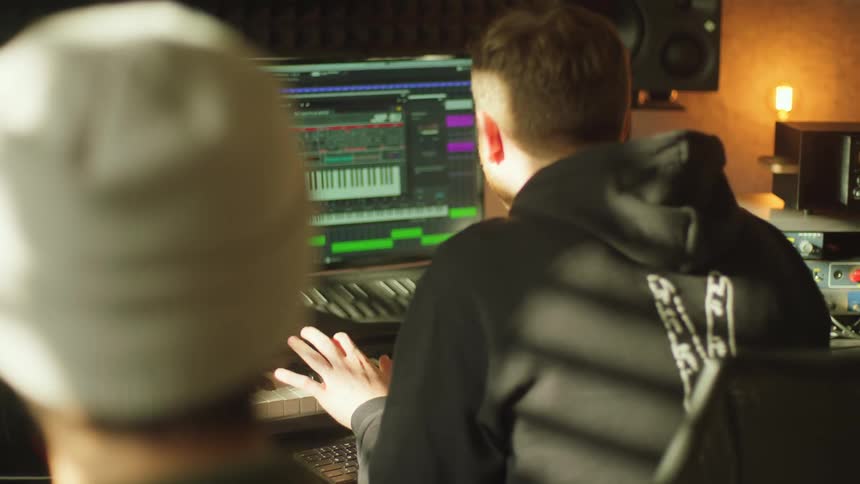Man with a black hoodie plays electric piano while man with a white beanie sings a song in the studio