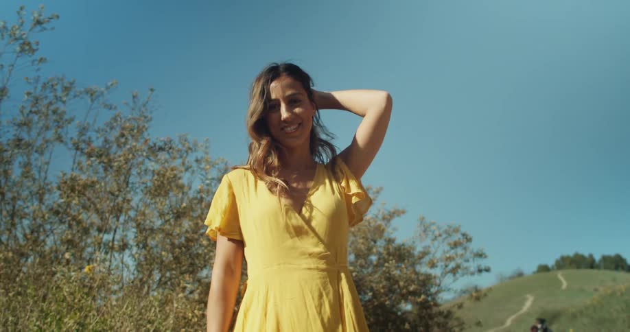 A woman in a yellow dress smiling and staring with plants trees the sky and clouds in the background