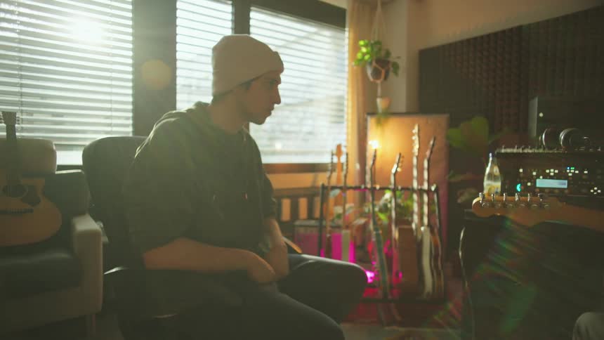 A man observes another man playing guitar in a room full of music equipment