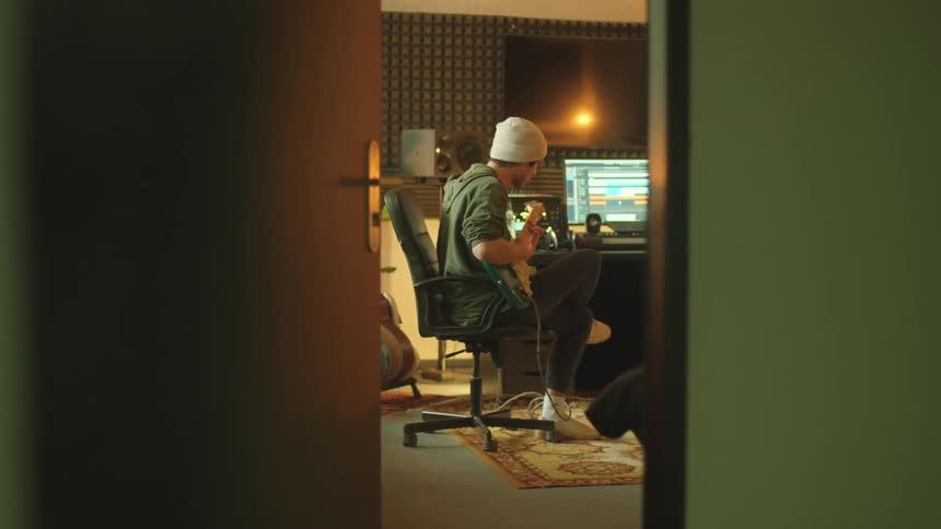 A man in a music studio sitting on a chair playing a guitar with a computer in the background