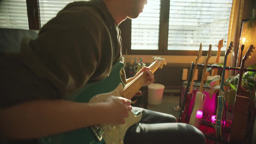 A man with a hat in a room playing an electric guitar with a wall and windows in the background