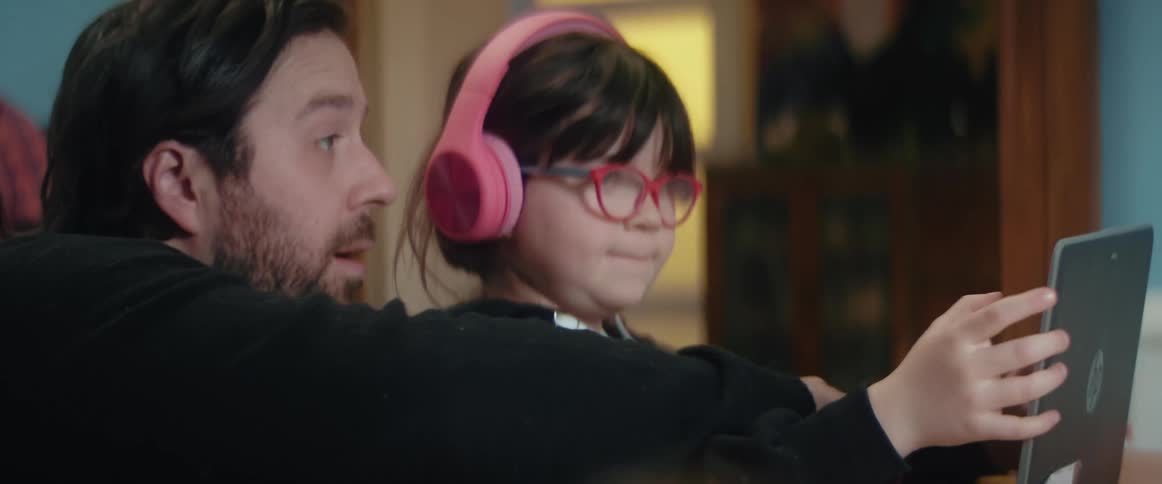 A man and a girl in a room with the girl using a laptop with headphones and glasses