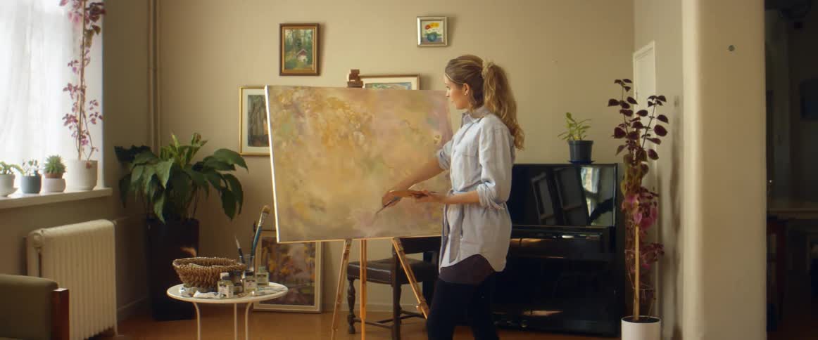 A woman holds a palette as she paints a canvas on an easel in her living room