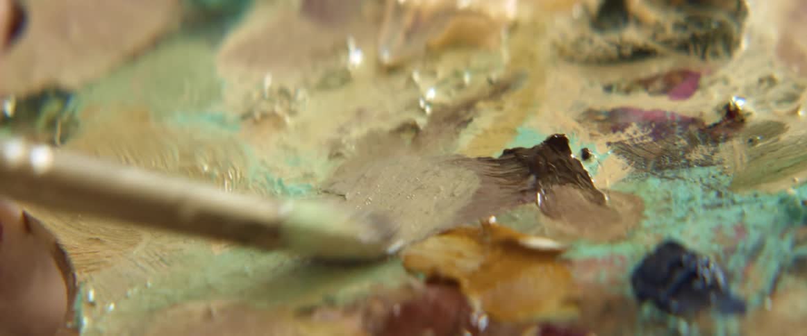 A painter rubs a paint brush in paint on a wooden palette