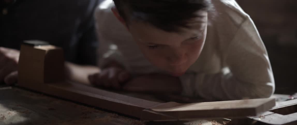 A boy blows wood shavings from a board