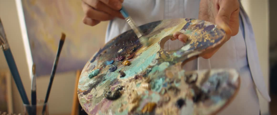 A person in a room brushing a paint brush on a painting pallet with a wall in the background