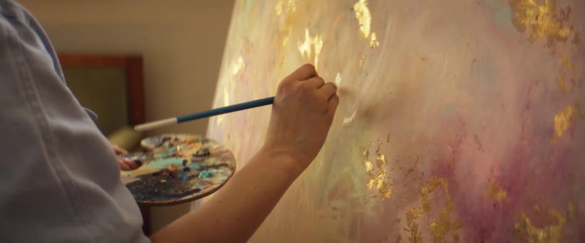 A painter paints on a large canvas while holding a palette covered in paint