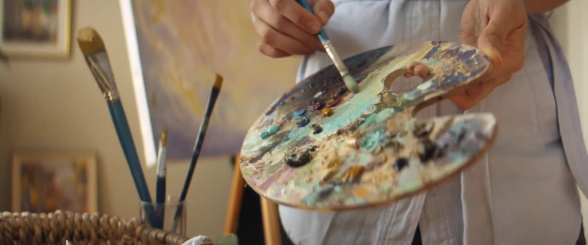 A person in a room adding paint to a painting pallet and mixing colors with a paint brush