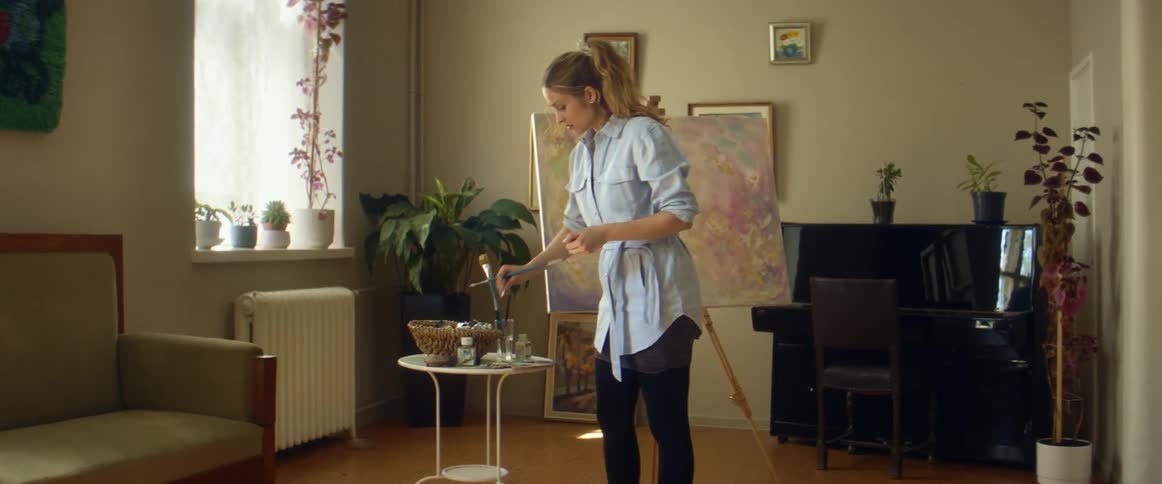 Blonde female painter with ponytail light blue shirt and black pants paints on a canvas next to plants and window in front of piano in room