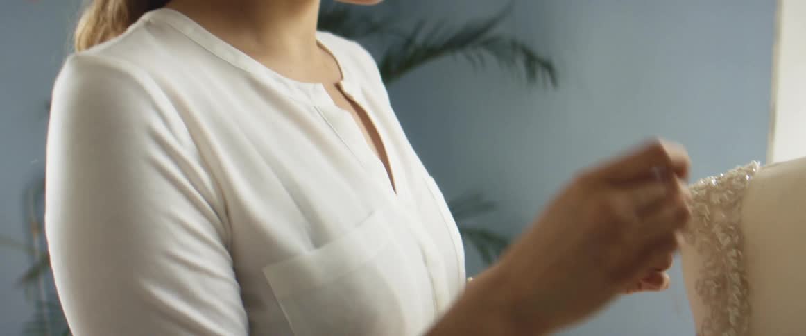 A woman in a room stitching a fabric with a plant a wall and window in the background