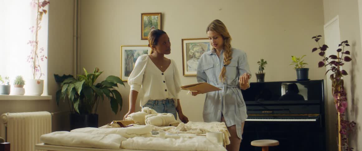 Two women in a room standing by a table with a piano plants and a wall in the background
