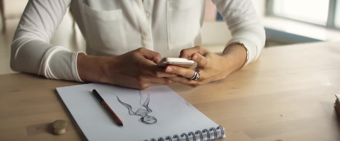 A woman sits at a table with a sketchbook and then picks up her phone