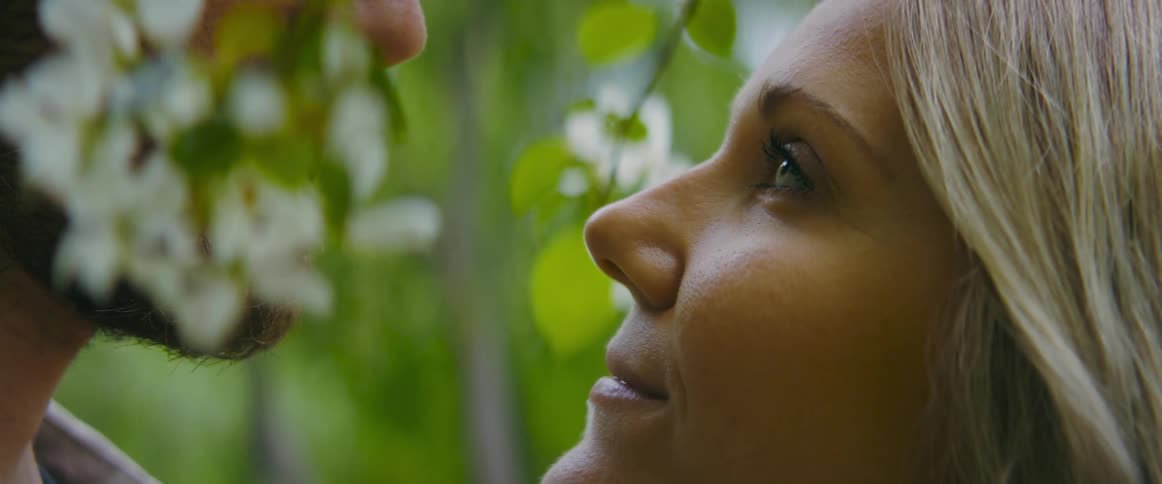 A woman and look gaze into one anothers eyes while standing beneath the blossoms flowers of a tree during the day