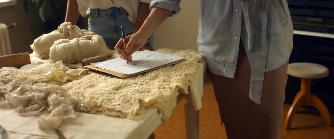 Two women stand at table covered in different fabrics as one sketches dress with piano in background