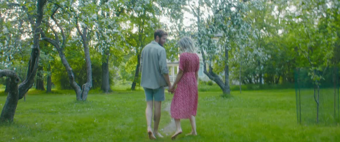 A man and woman hold hands and stop to kiss as they walk barefoot in the grass