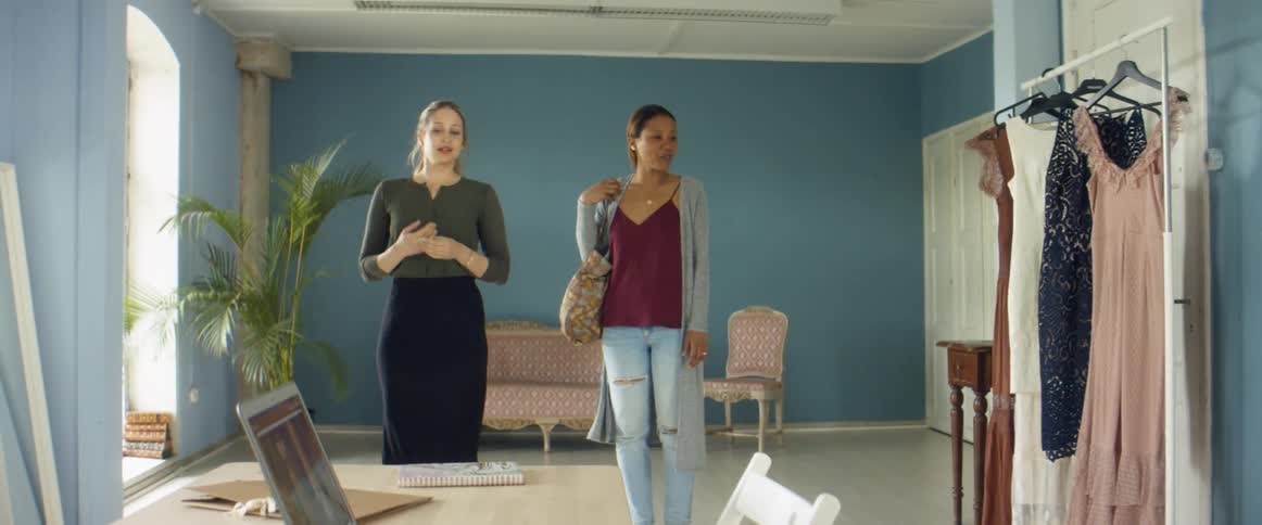 Two women talk as they enter office with dresses hung on rack