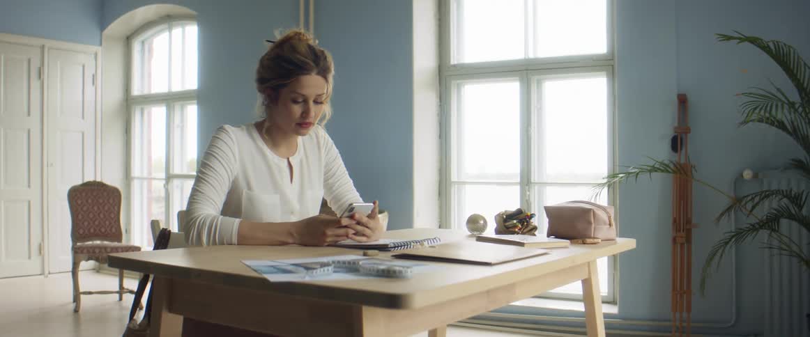 A woman sits at her desk and picks up her cell phone and looks at it