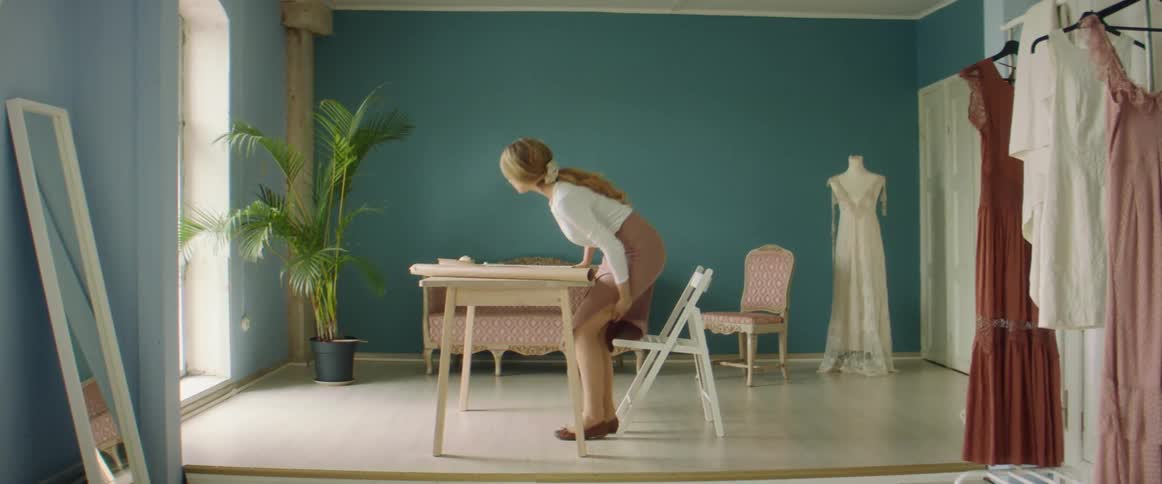 A fashion designer carrying rolls of paper approaches and sits at a desk in front of a large window