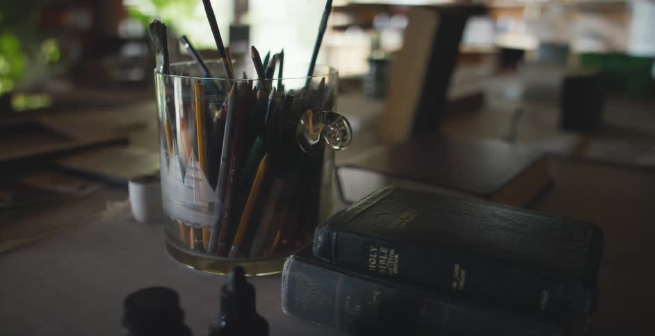 Leather bound books including a bible sit on a desk next to a glass container full of pens pencils and paint brushes