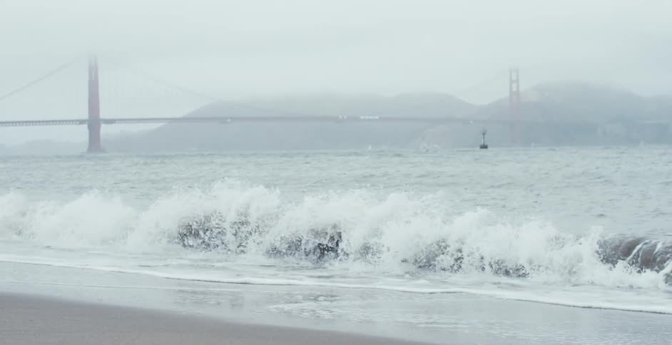 Waves crashing on the sands of a beach next to a suspension bridge covered in fog