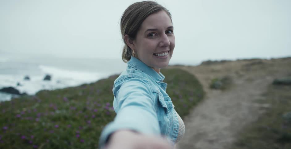 A woman turns around and smiles as she reaches her hand out while walking toward the edge of a cliff