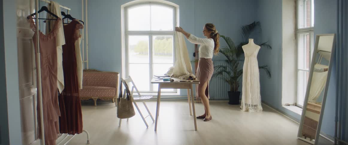 Fashion designer stands next to wooden table examining cloth with dress form in corner and dresses hanging on clothing stand
