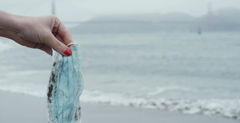 A woman holds a dirty mask on the beach as the waves roll in and seagulls fly