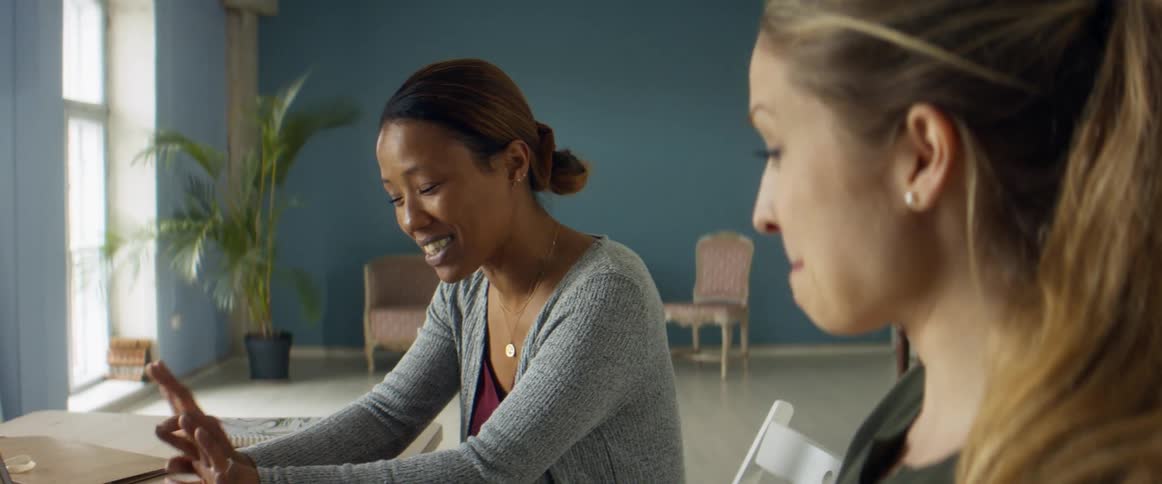Two women sit at a table and talk about the laptop in front of them