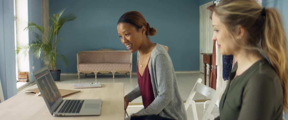 Two women in a room sitting at a desk and looking at a laptop with a wall and window in the background