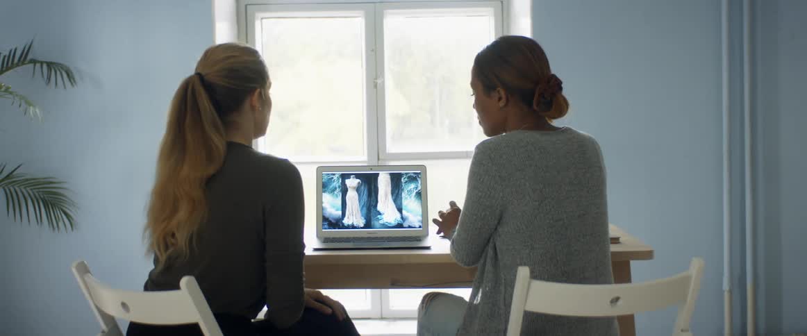 Two women discuss as they are sitting at a desk and looking at a dress on laptop screen