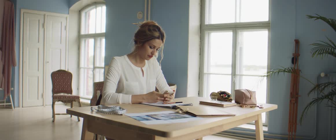 A woman in a room sitting at a table with a notepad using a smart phone