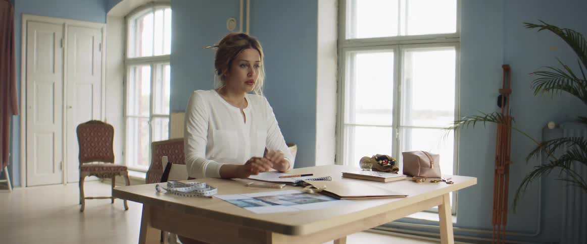 A woman in a room sitting at a table using a smart phone with a wall and windows in the background