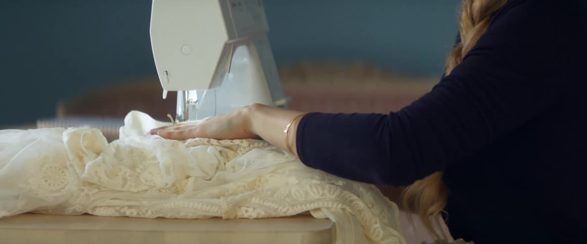A woman in a room sewing a fabric with a sewing machine with a wall in the background