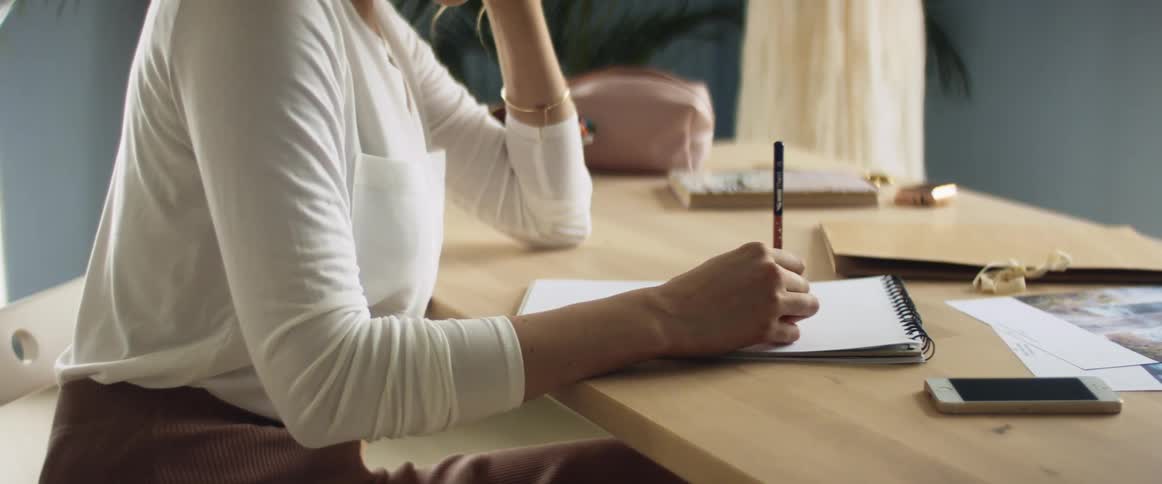 A dress designer sits at a table and draws a design in her notebook