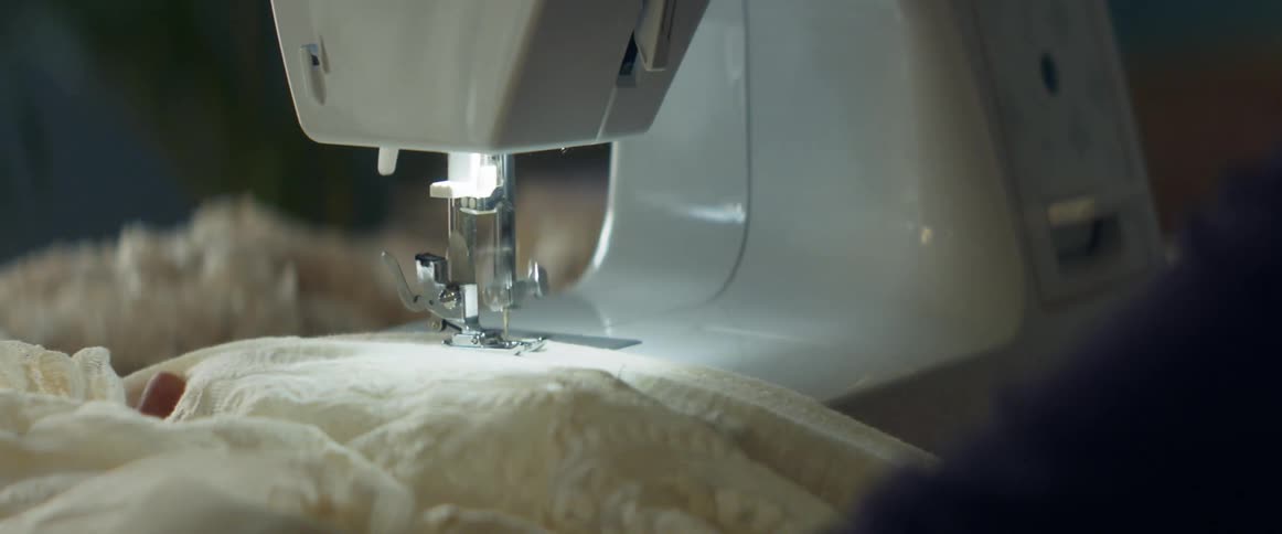 A sewing machine stitches a hem on an off white lace fabric