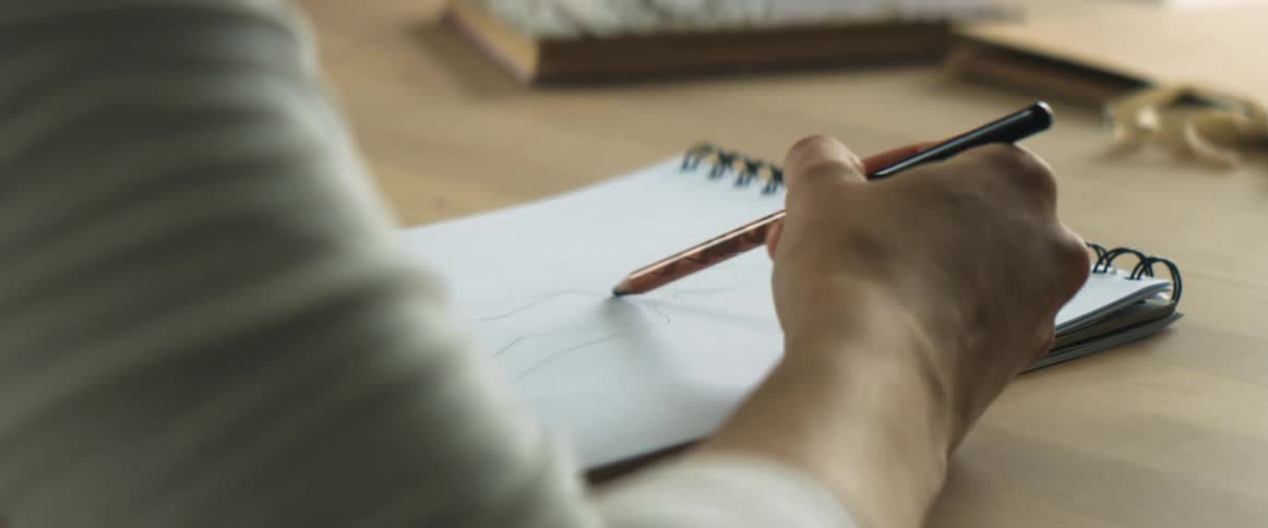 Woman sits sketching in sketchpad set on wooden surface