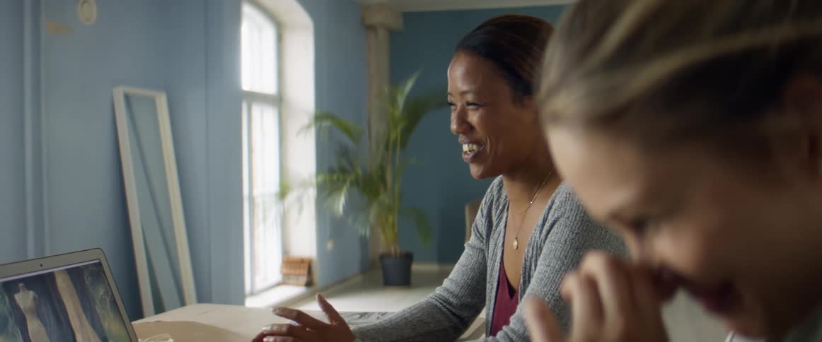 Two women sit beside each other at a table and a laptop laughing together