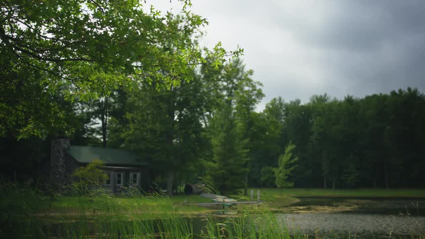 A cabin in the woods next to a pond with trees the sky and clouds in the background